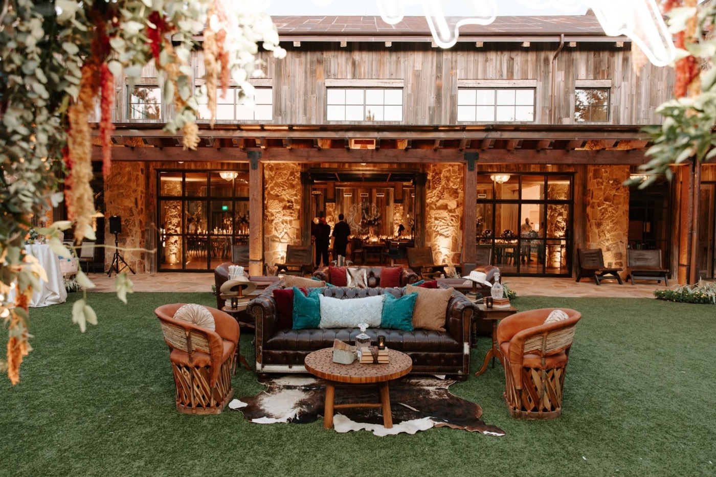 An outdoor wedding lounge styled with leather seating, pillows, and hanging florals at a rustic venue