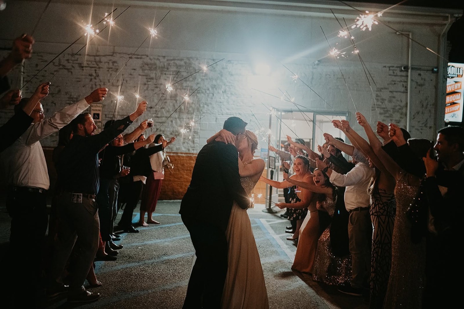 A bride and groom kissing during a sparkler send-off surrounded by guests at night